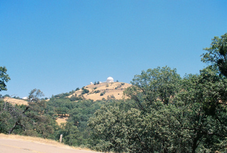 [At the top of a dirt mound is a white domed structure with two smaller white structures to the left of it. There are trees at the roadside which is at the bottom of the image. There are also trees partway up the hillside, but only a few at the top.]